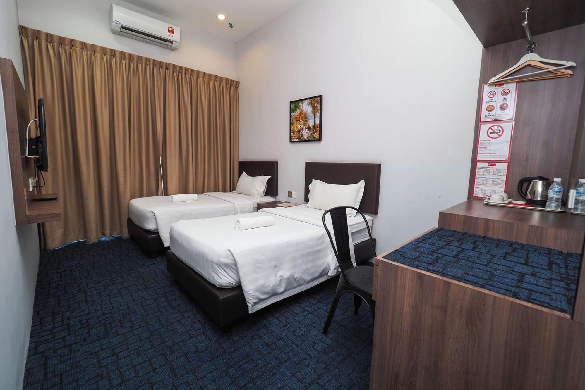 Habitación de hotel con dos camas, un escritorio, una silla, aire acondicionado, un cuadro y un cartel con normas en la pared.