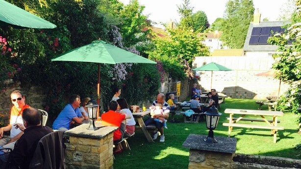 An outdoor patio area with stone tables and green umbrellas, surrounded by lush greenery and stone walls. - The Brewers Arms (South Petherton)
