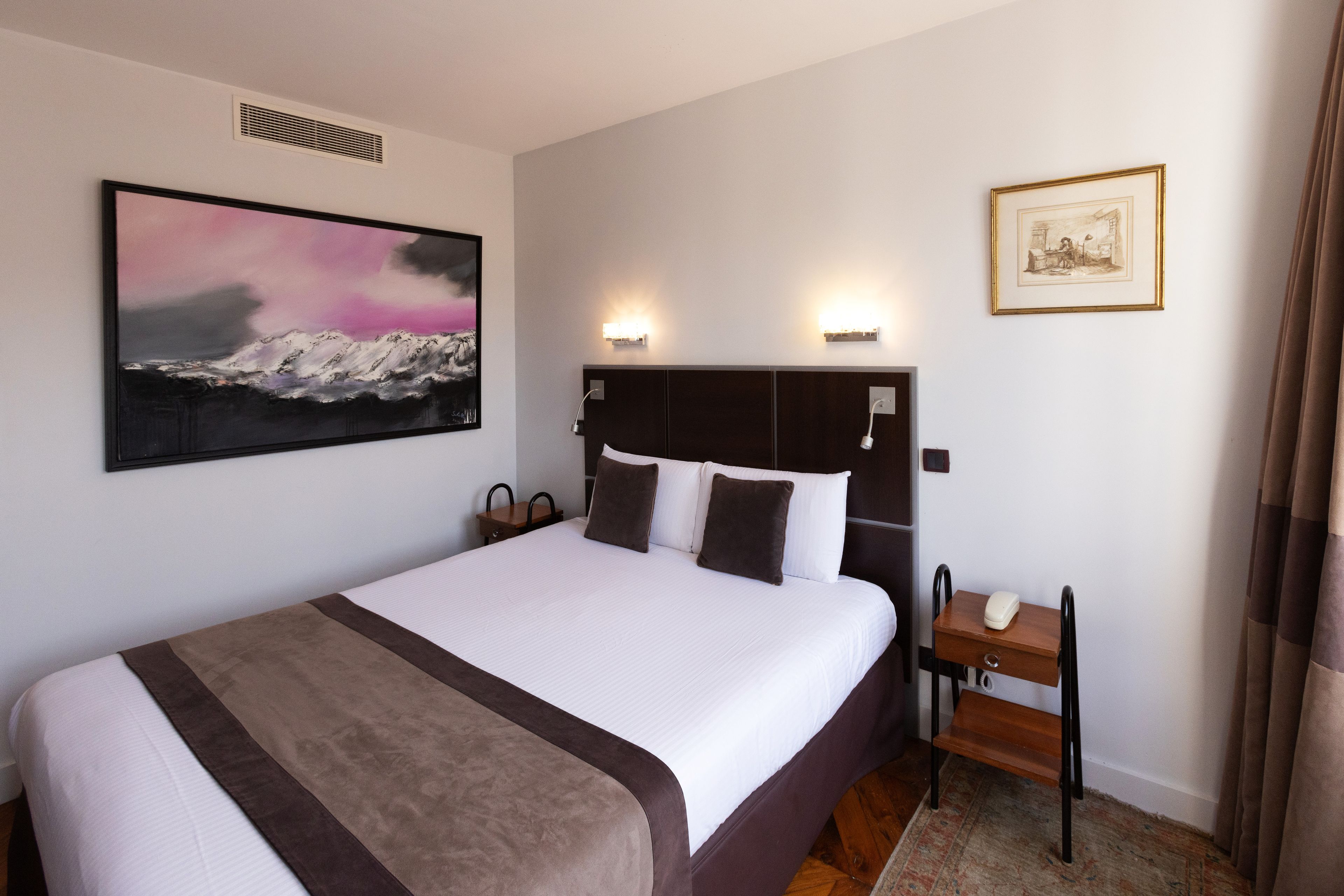A hotel room with a large bed, a bedside table, a nightstand, a wall-mounted lamp, a painting, and a framed picture.