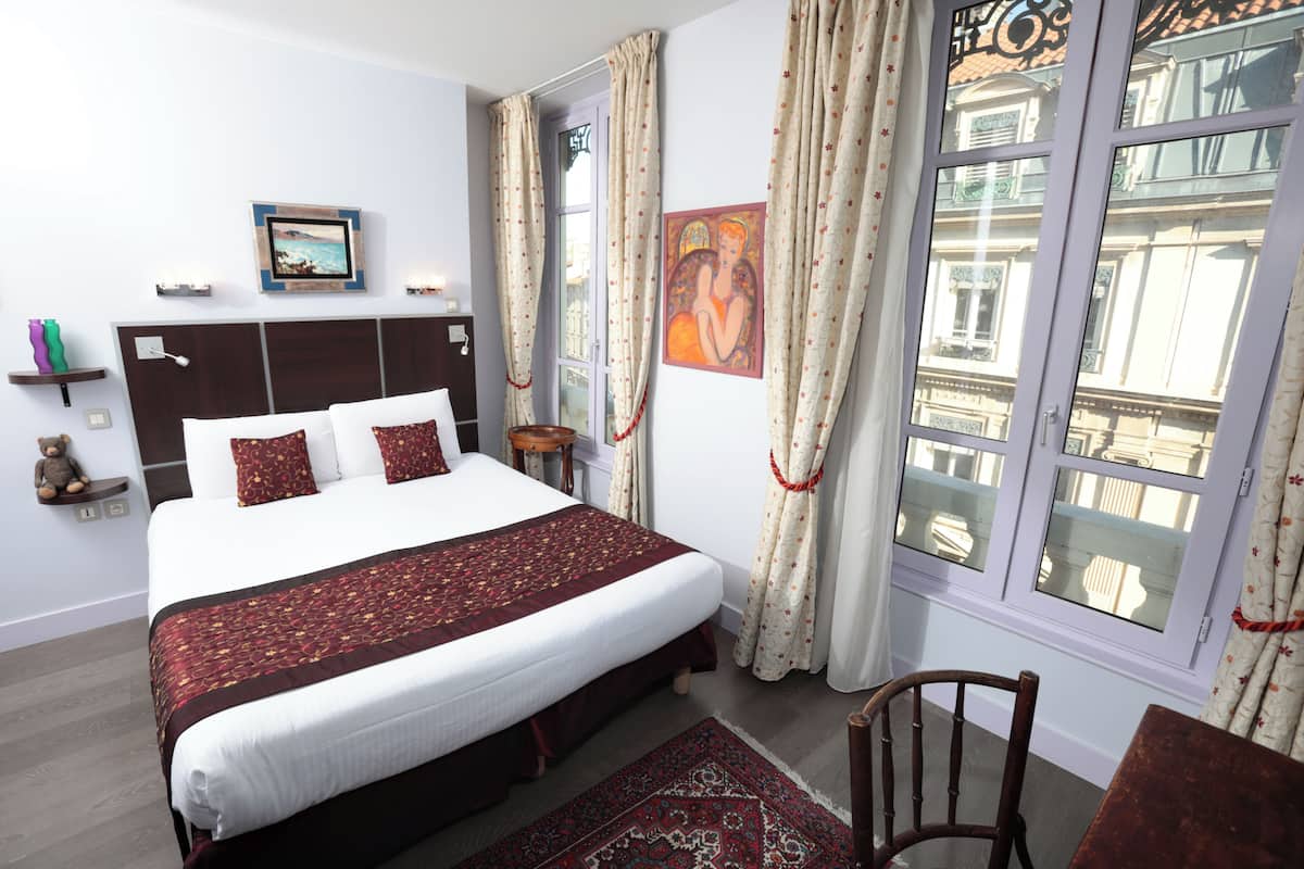 Une chambre à coucher bien rangée, avec un grand lit, une chaise en bois, une petite table, une table de chevet et une fenêtre avec des rideaux.