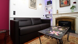 Uma sala de estar com um sofá preto, uma mesa com azulejos florais, uma lareira e prateleiras fixadas na parede com livros e objetos decorativos.