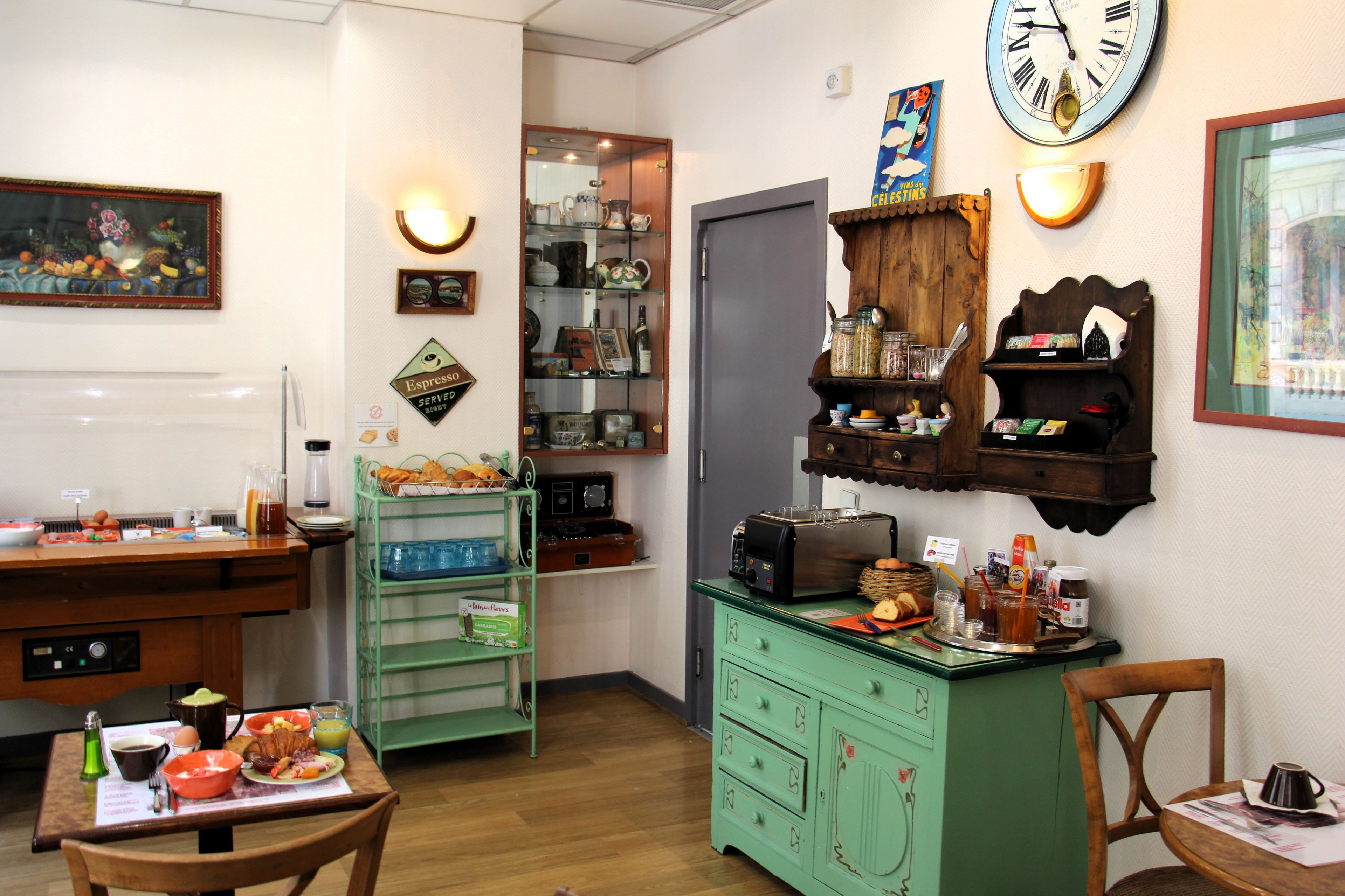 Une pièce avec une table à manger dressée pour le petit-déjeuner, un buffet vert avec des tiroirs, une horloge au mur et des étagères remplies de divers objets.