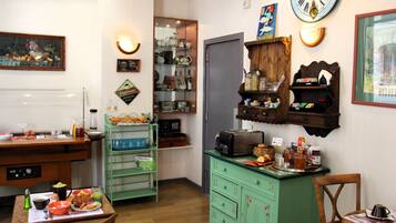 Une pièce avec une table à manger dressée pour le petit-déjeuner, un buffet vert avec des tiroirs, une horloge au mur et des étagères remplies de divers objets.