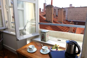Un balcone con vista su tetti rossi in cotto, un tavolo di legno con due tazze e piattini, una scatola con l'etichetta 'Cacao Suchard' e un bollitore.