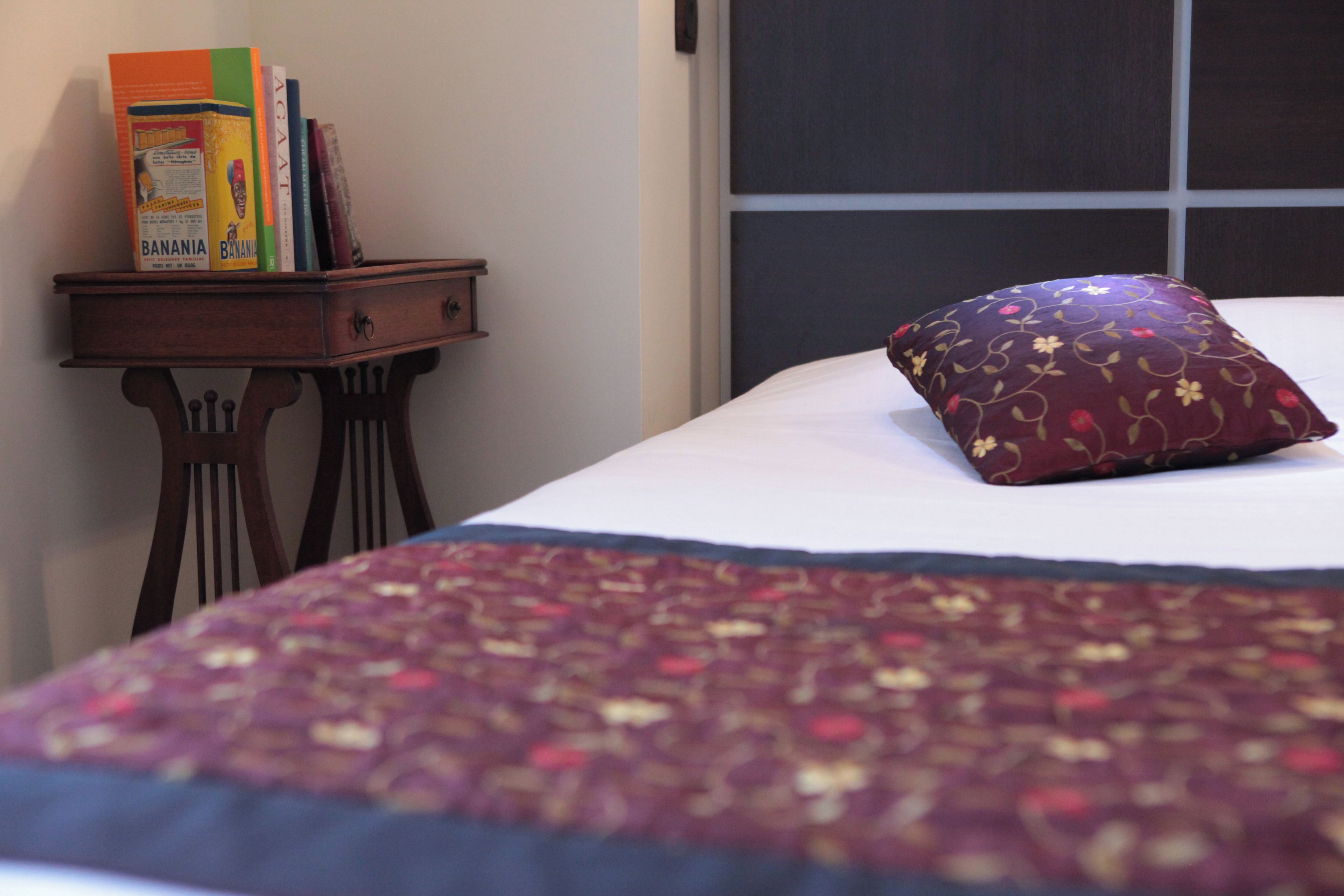 A bed with a patterned bedspread and a decorative pillow, a nightstand with books and a box, and a plain wall.