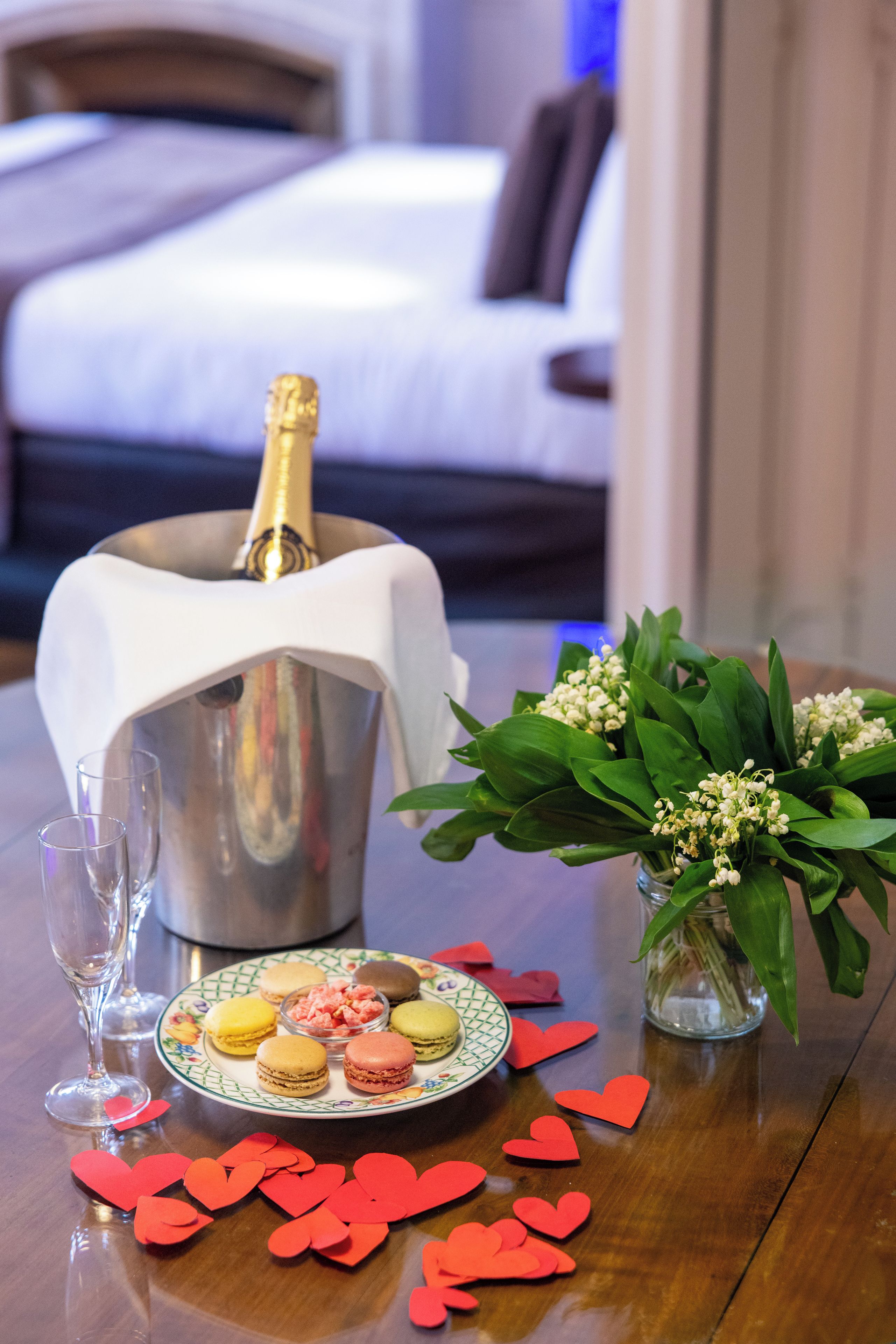 Ein Tisch mit einer Champagnerflasche, einer Macaron-Platte, einer Blumenvase und verstreuten, herzförmigen roten Objekten.