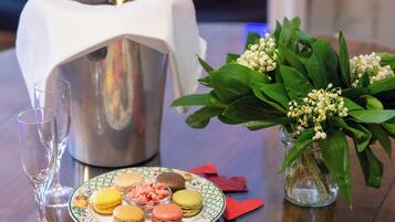 Ein Tisch mit einer Champagnerflasche, einer Macaron-Platte, einer Blumenvase und verstreuten, herzförmigen roten Objekten.