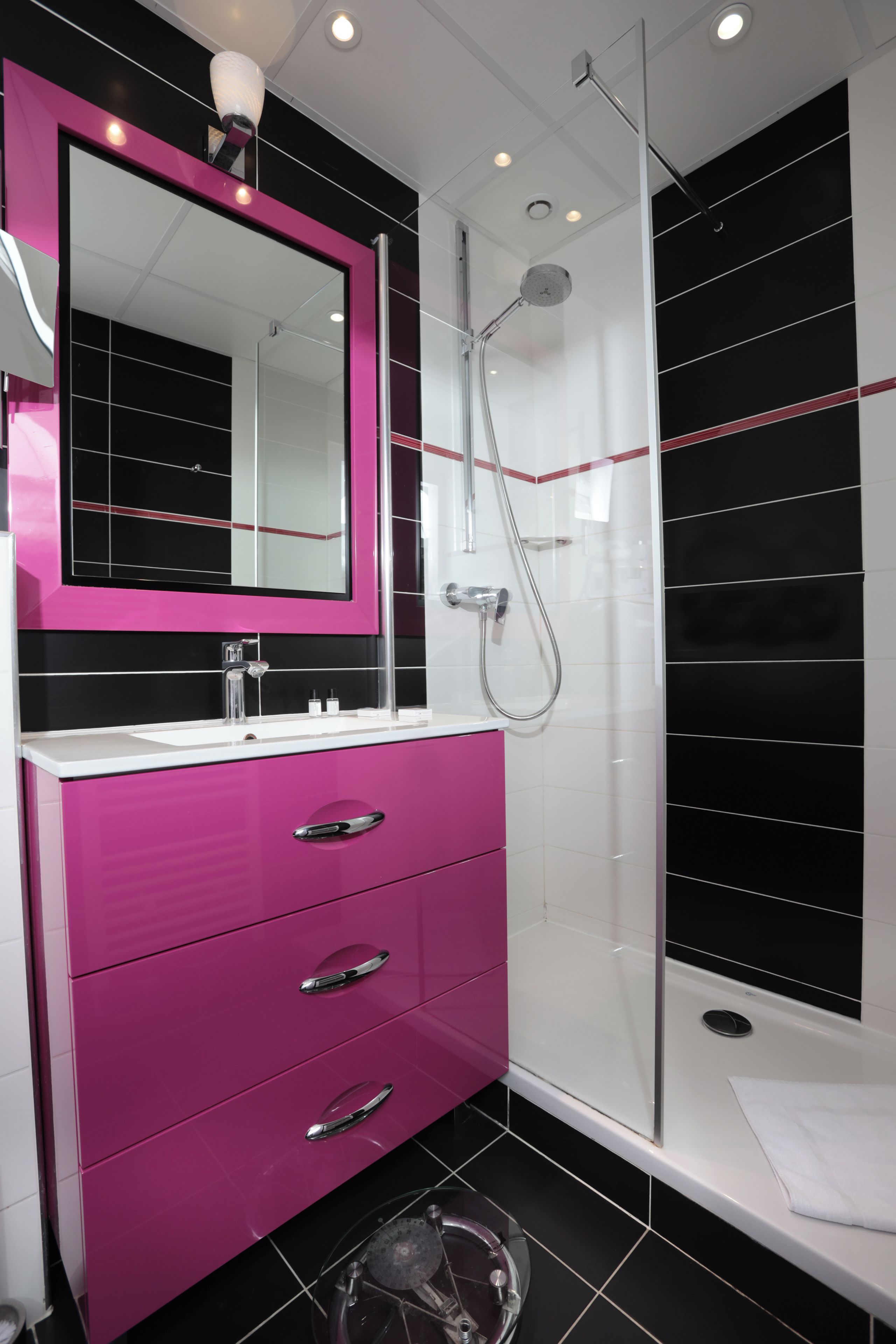 Un baño moderno con cabina de ducha de vidrio, un tocador rosa con espejo y paredes con azulejos blancos y negros.