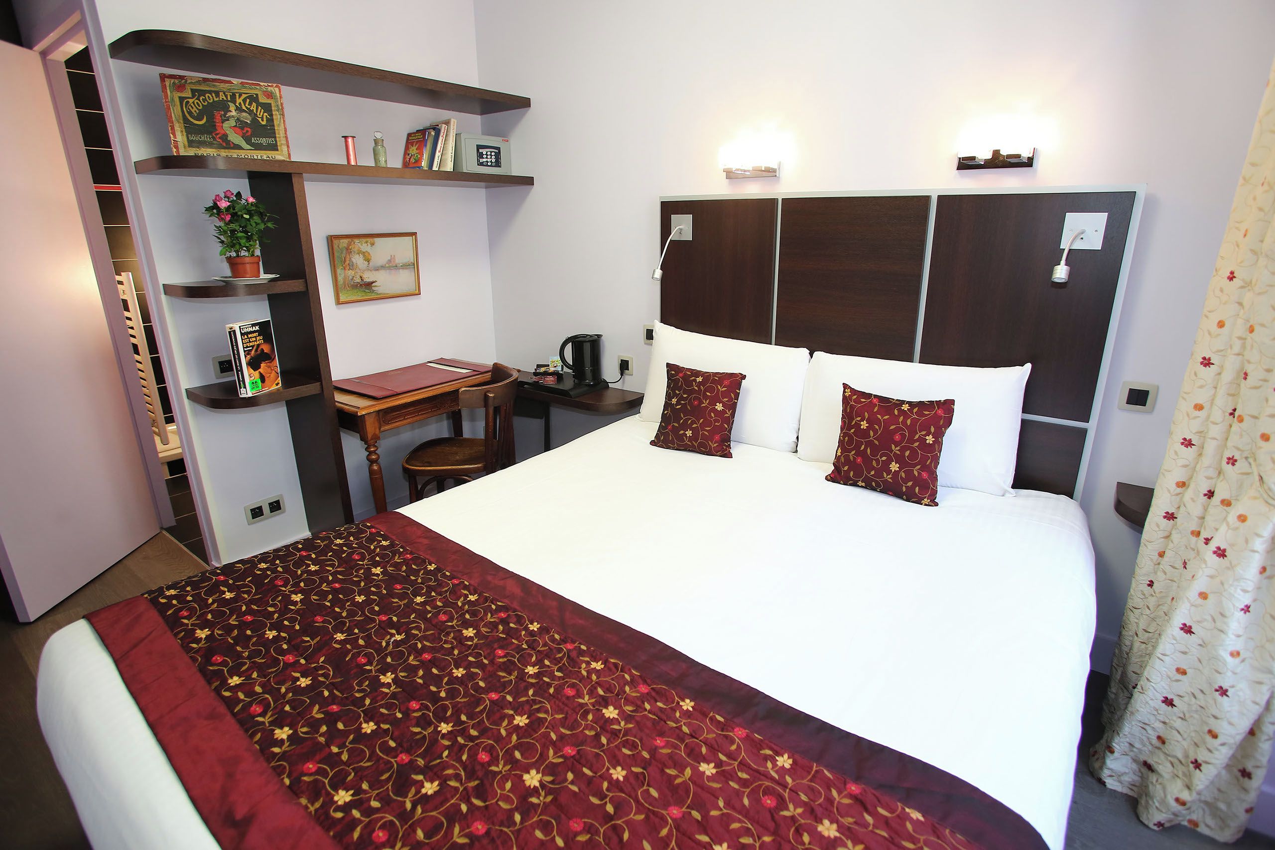Una habitación de hotel con una cama, un escritorio, una silla, una estantería y un cuadro.