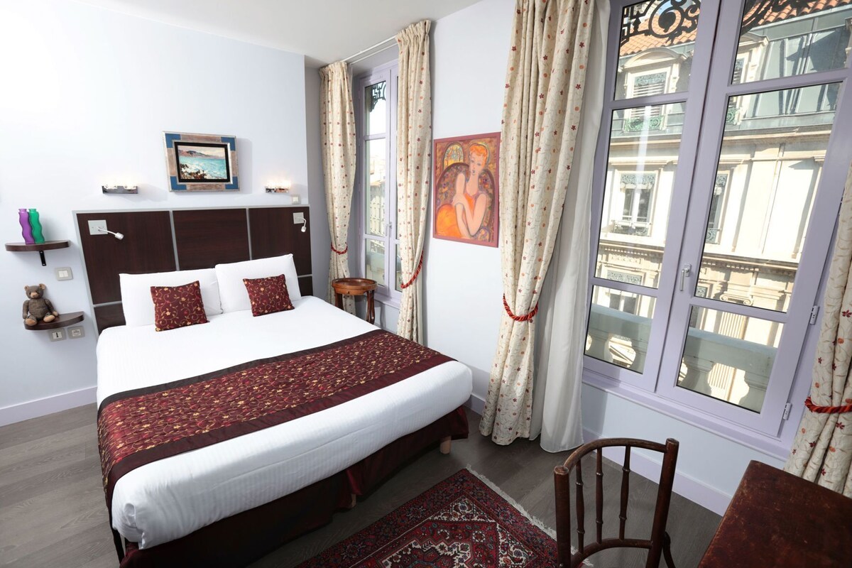A hotel room with a large bed, a wooden headboard, a nightstand, a chair, a window with curtains, and a view of buildings outside.