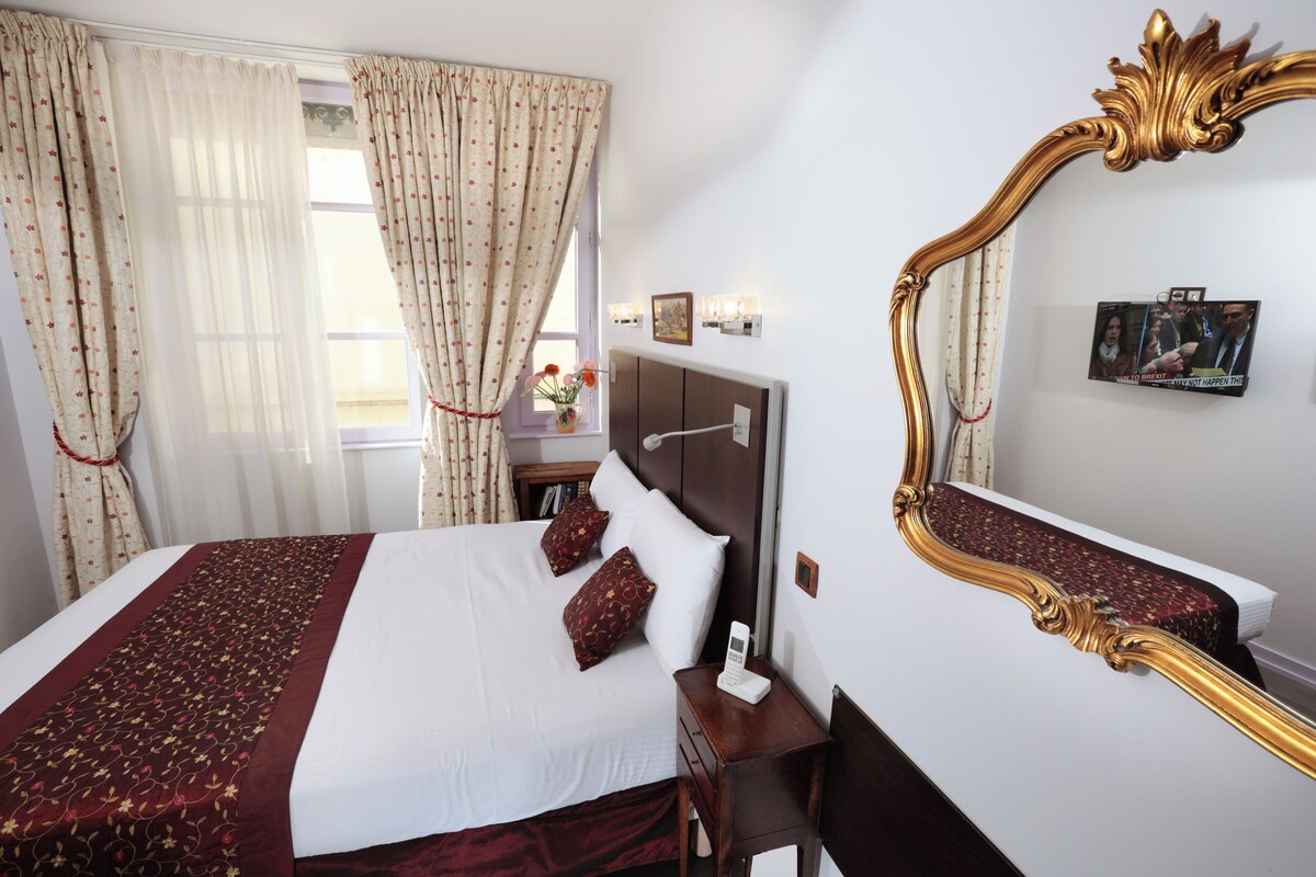 A neatly made bed with a patterned bedspread, a bedside table with a phone, and a large ornate mirror on the wall.