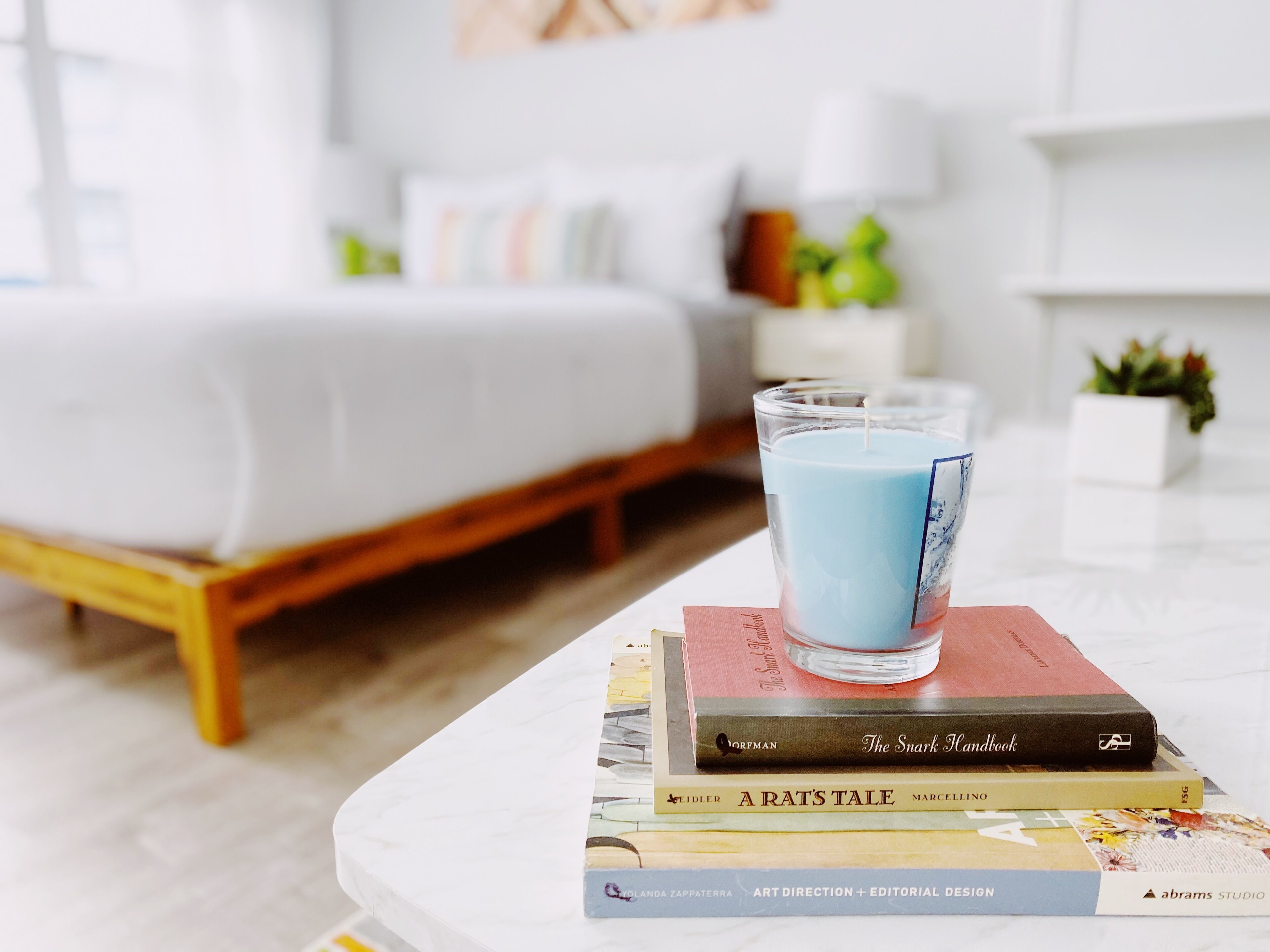 A neatly arranged stack of books on a white table with a clear glass candle holder containing a blue candle.