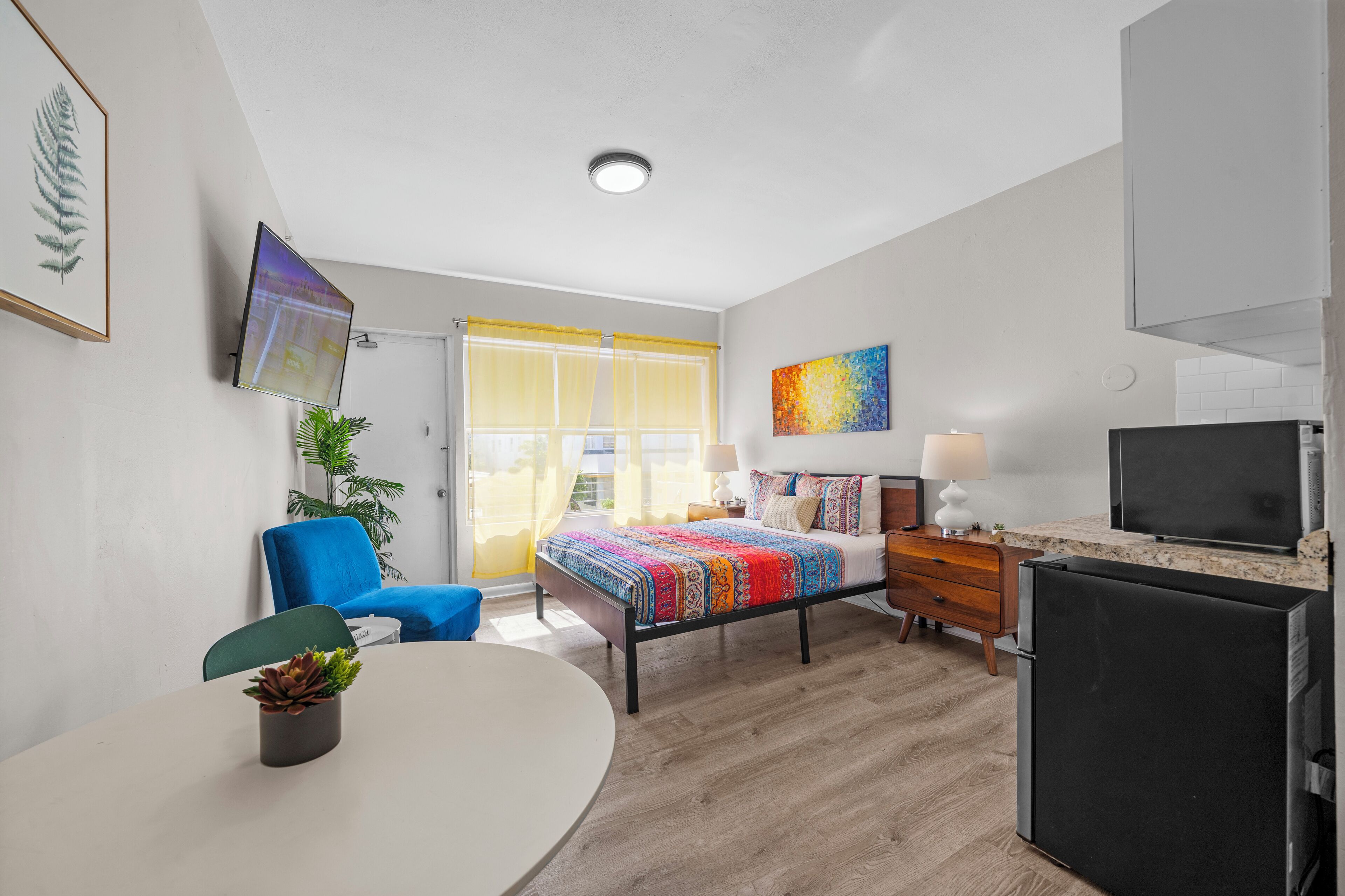 A modern hotel room with a bed, a desk, a chair, a TV, and a refrigerator.