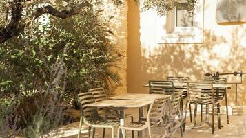 An outdoor seating area with tables and chairs, surrounded by greenery and a building in the background.