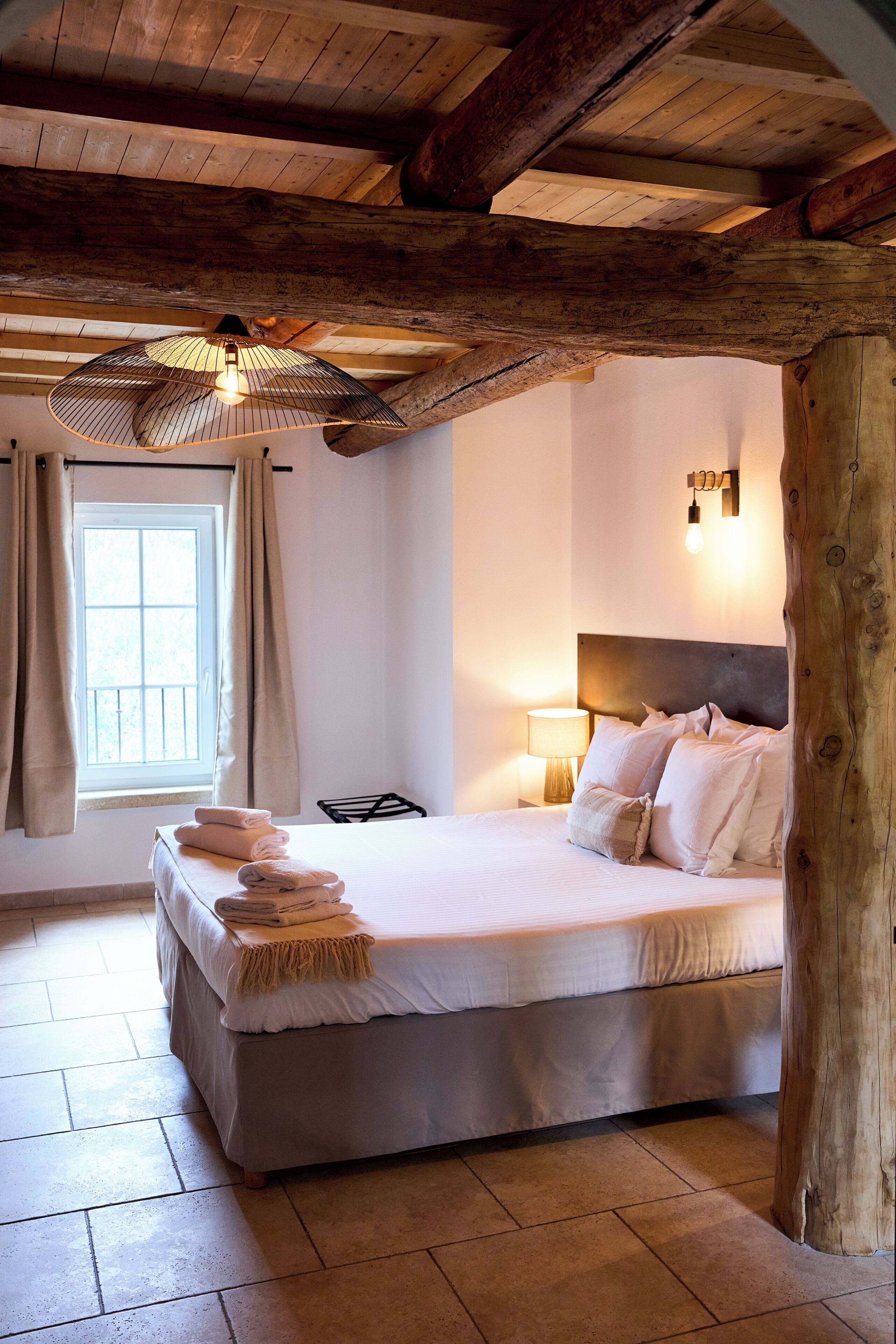 Une chambre avec un plafond en bois, un lit recouvert de draps blancs, une lampe de chevet et une fenêtre avec des rideaux.