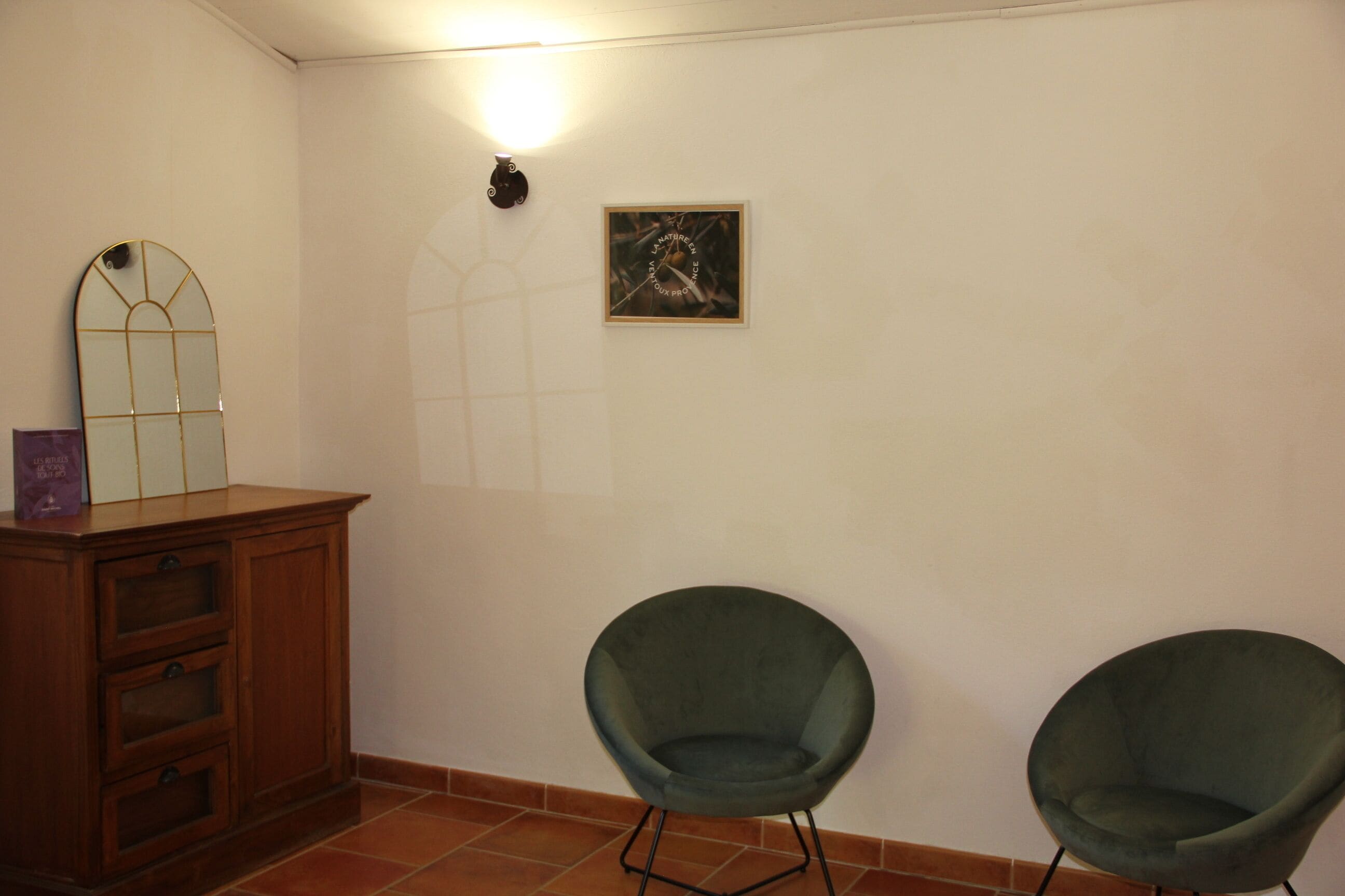 A room with a wooden cabinet, two green chairs, a wall-mounted light, and a framed picture.