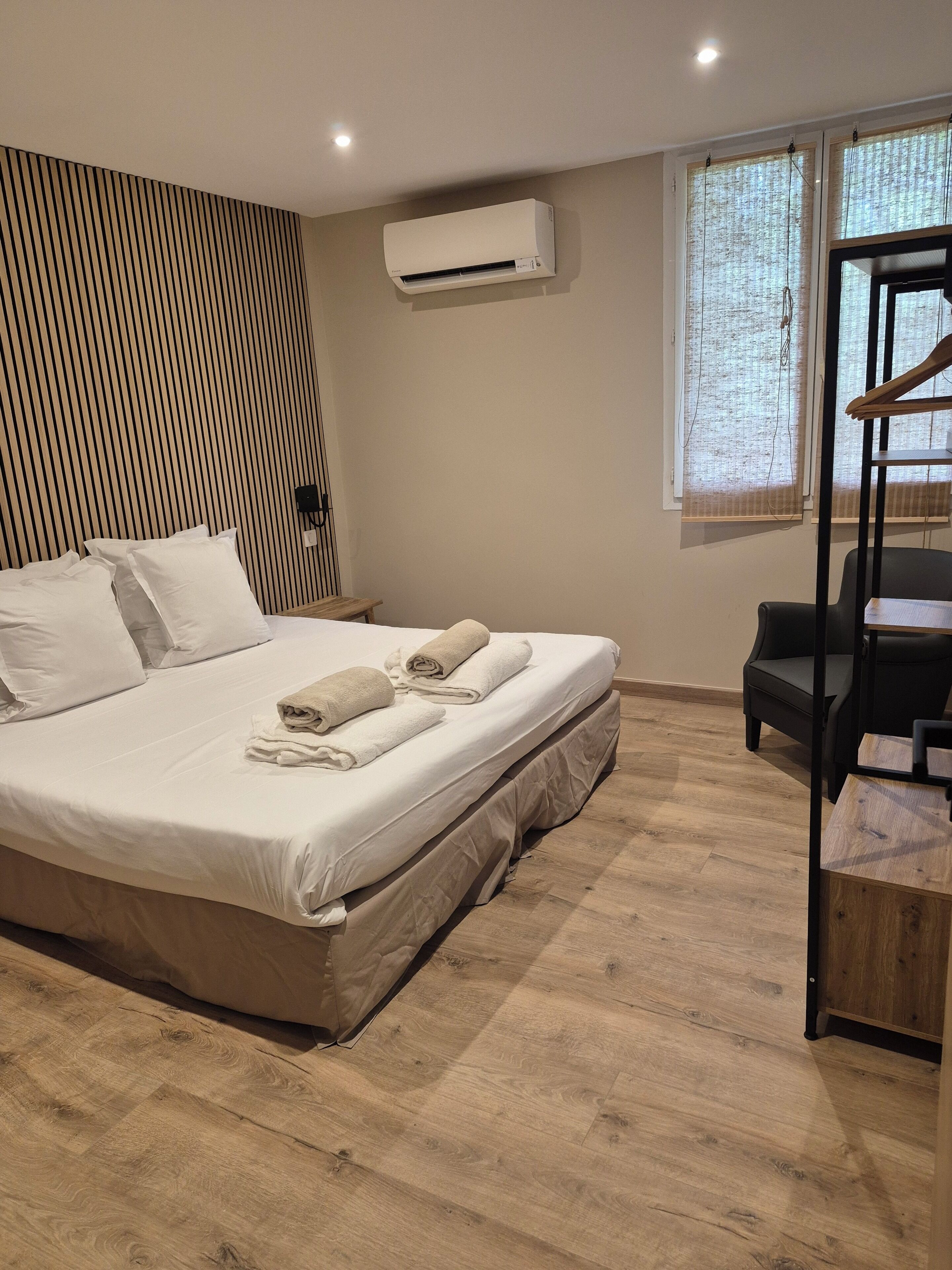 A neatly made bed with white linens and rolled towels, a wooden floor, a black chair, a small wooden table, and a wall-mounted air conditioner.