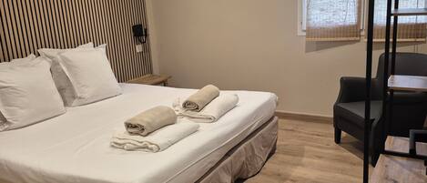 Una cama bien hecha con sábanas blancas y toallas enrolladas, piso de madera, una silla negra, una mesita de madera y un aire acondicionado de pared.