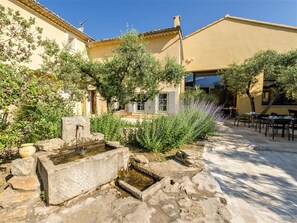 A courtyard with a stone fountain, outdoor seating, and a building with a yellow facade. - Le Clos Saint-Michel & Spa (Malaucene)