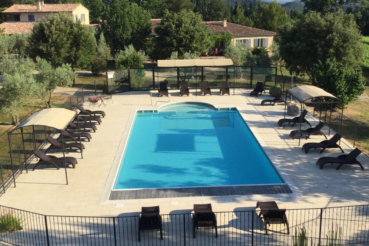 A rectangular swimming pool with a tiled deck, surrounded by lounge chairs and sunbeds, with a fence and trees in the background.