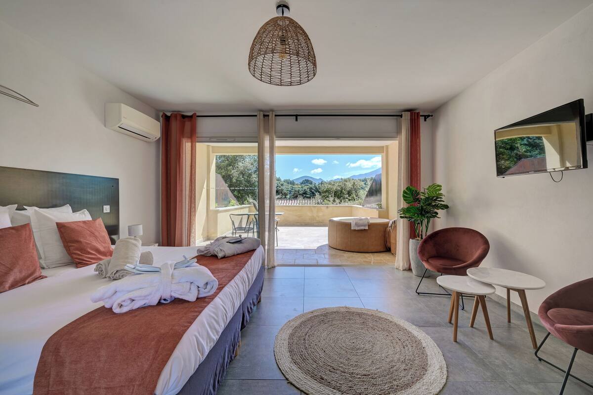 A modern hotel room with a large bed, a seating area with two chairs, a small table, and a large window offering a view of the outdoors.