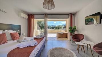 A modern hotel room with a large bed, a seating area with two chairs, a small table, and a large window offering a view of the outdoors.
