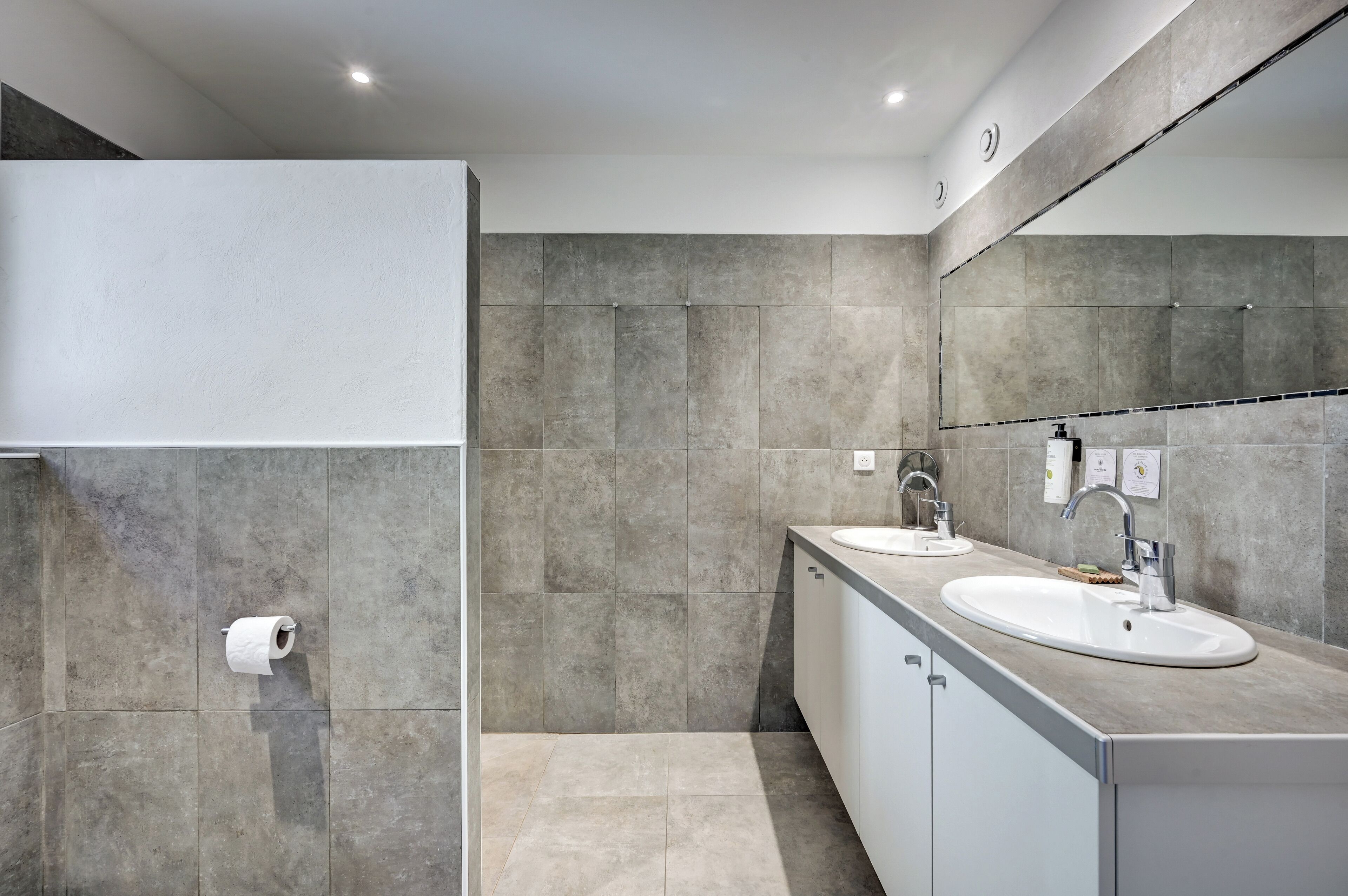A modern bathroom with two sinks, a large mirror, and a toilet paper holder on the wall.