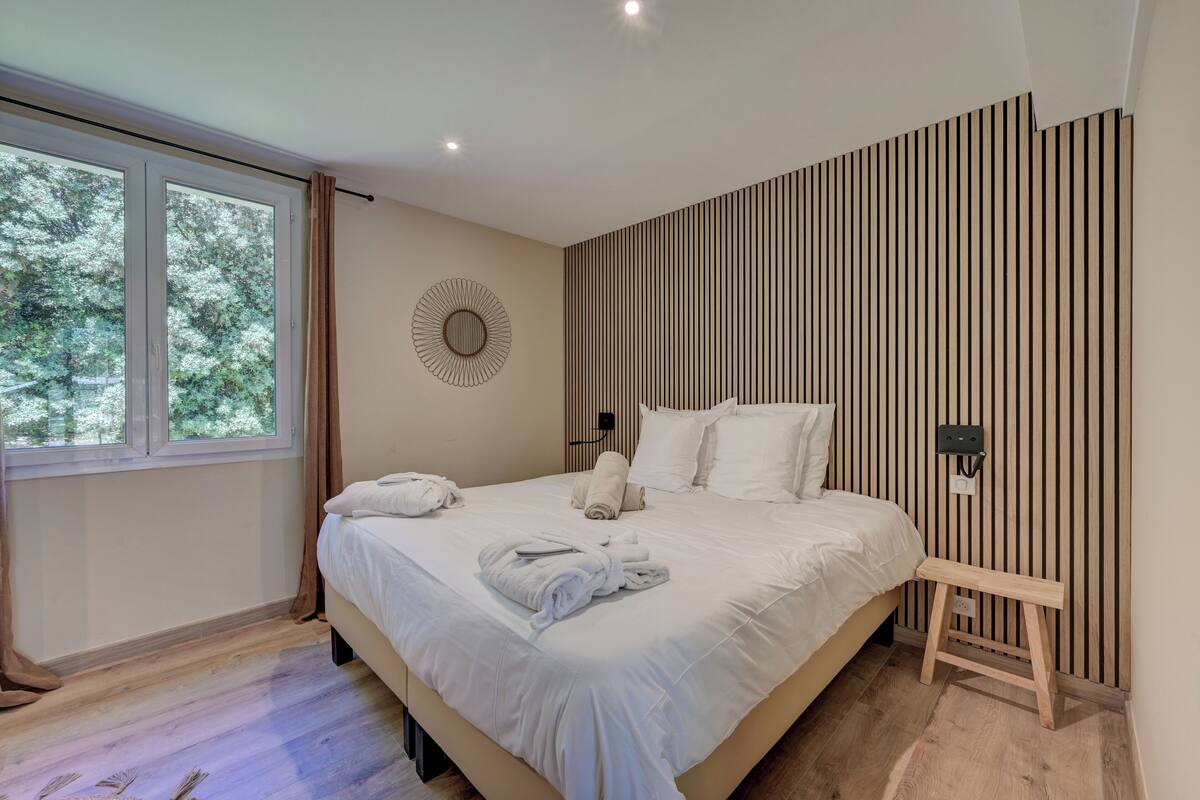 A modern bedroom with a large bed, a wooden bench, a wall-mounted light, and a window with curtains.