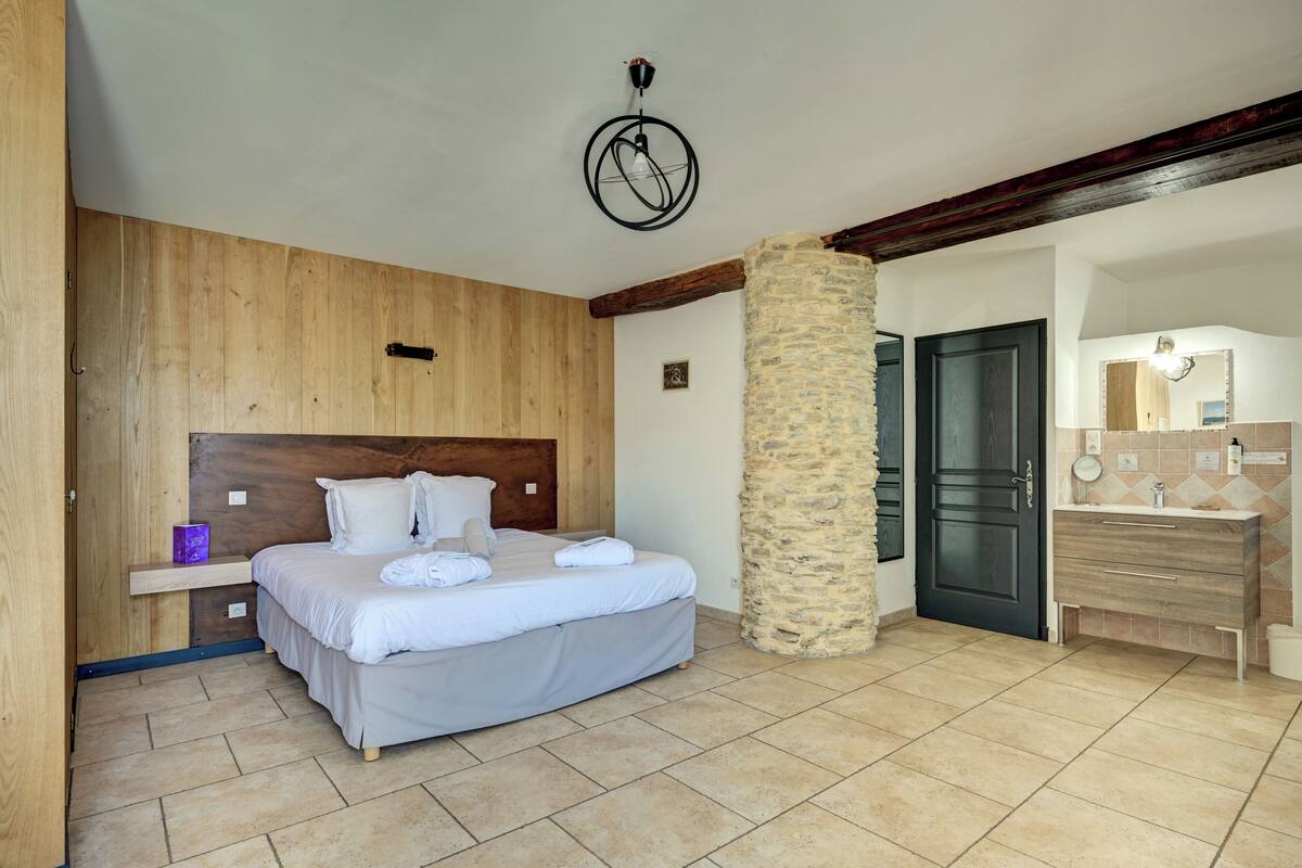 A bedroom with a wooden headboard, a stone pillar, a wooden dresser, and a tiled floor.
