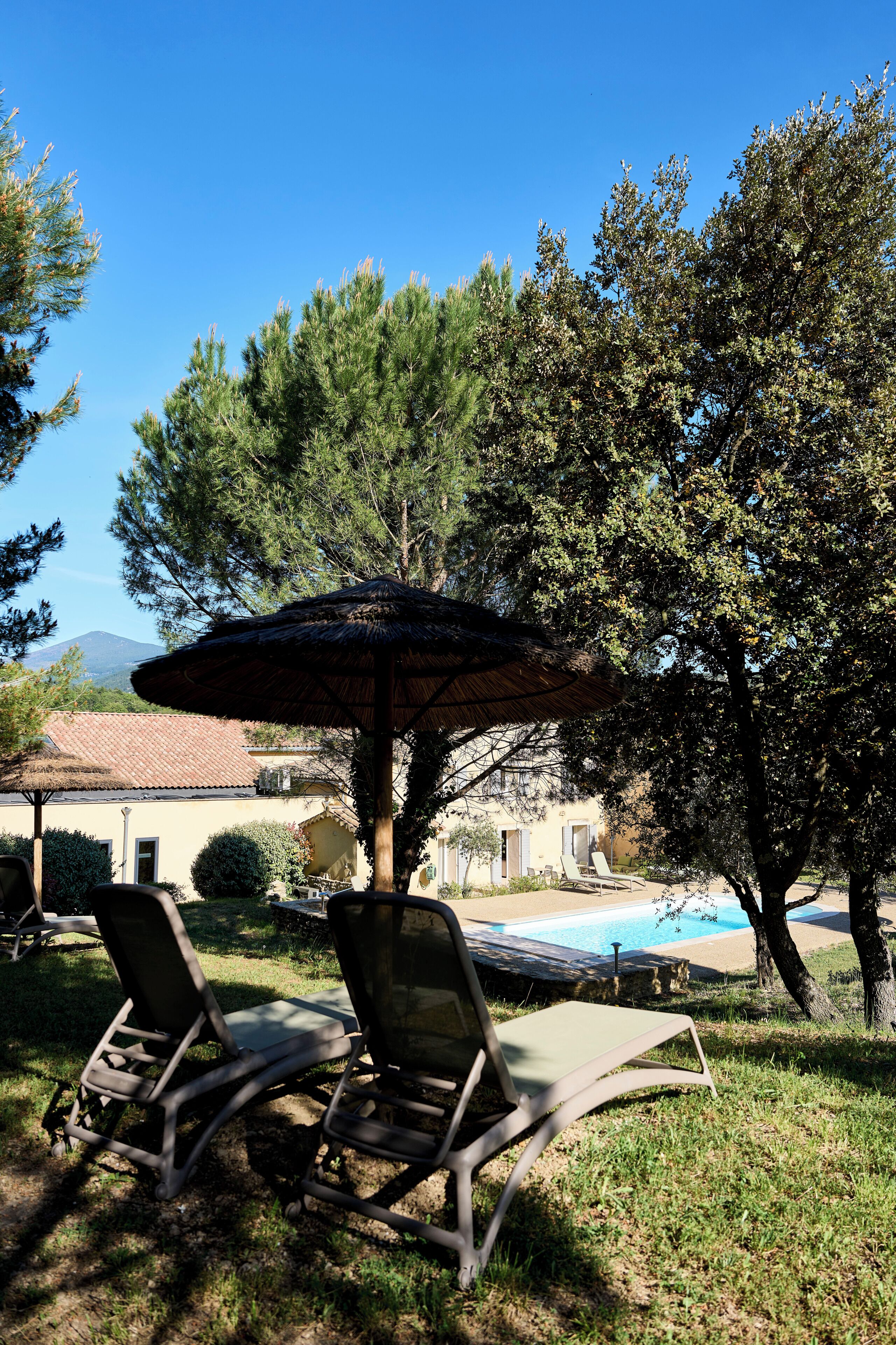 A poolside area with lounge chairs and a thatched umbrella, surrounded by trees and a building with a tiled roof.