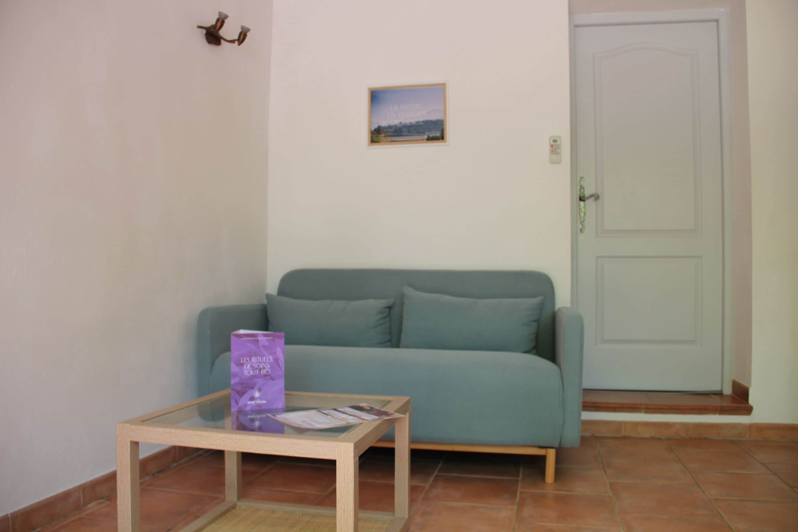 A room with a teal sofa, a small glass table with a purple bag, a framed picture on the wall, and a white door.