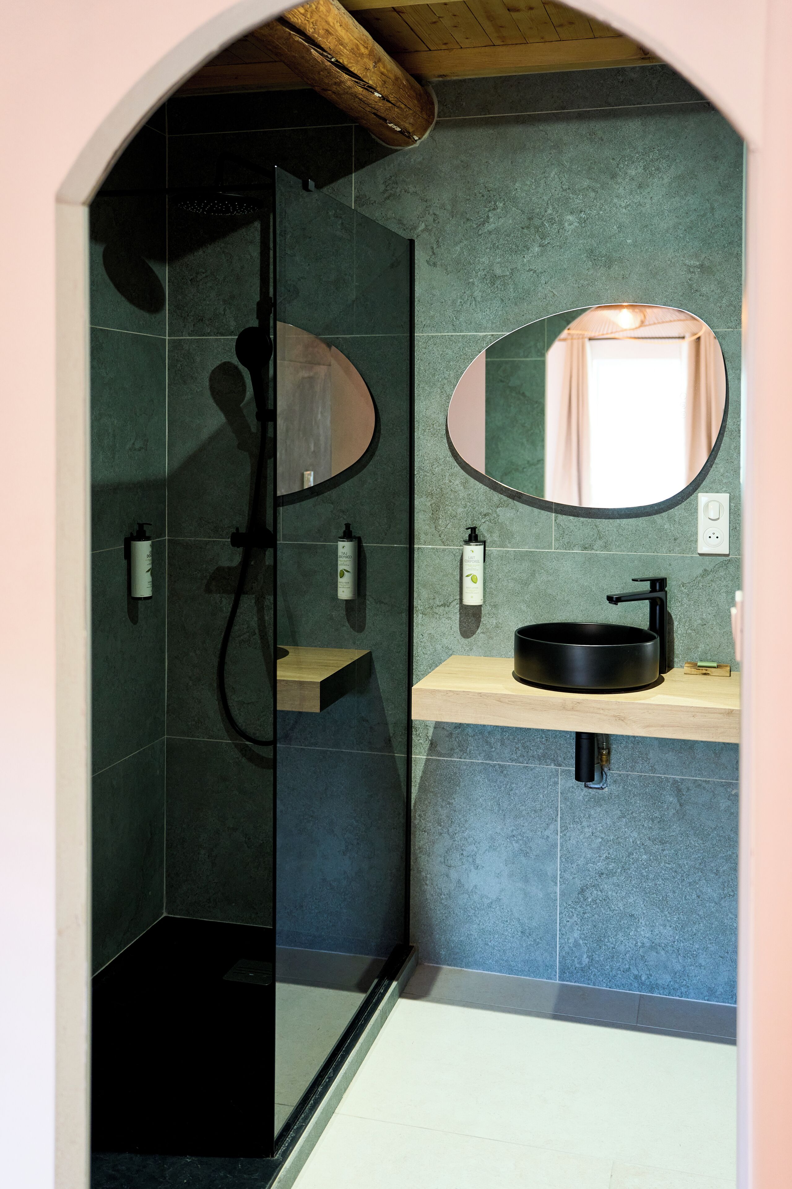 A modern bathroom with a glass shower enclosure, a wooden ceiling, a round mirror, and a black sink.