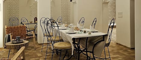 Un comedor con piso a cuadros, paredes blancas y arcos ornamentales. Hay mesas dispuestas con manteles blancos y sillas de metal negro.