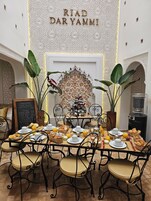 Un comedor con una mesa lista para el desayuno, destacando una pared decorativa con la inscripción "RIAD DAR YAMMI" y un árbol de Navidad.