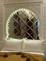Una cama con almohadas blancas y una manta marrón, frente a una pared con diseños blancos elaborados y un espejo grande con marco de diamantes.