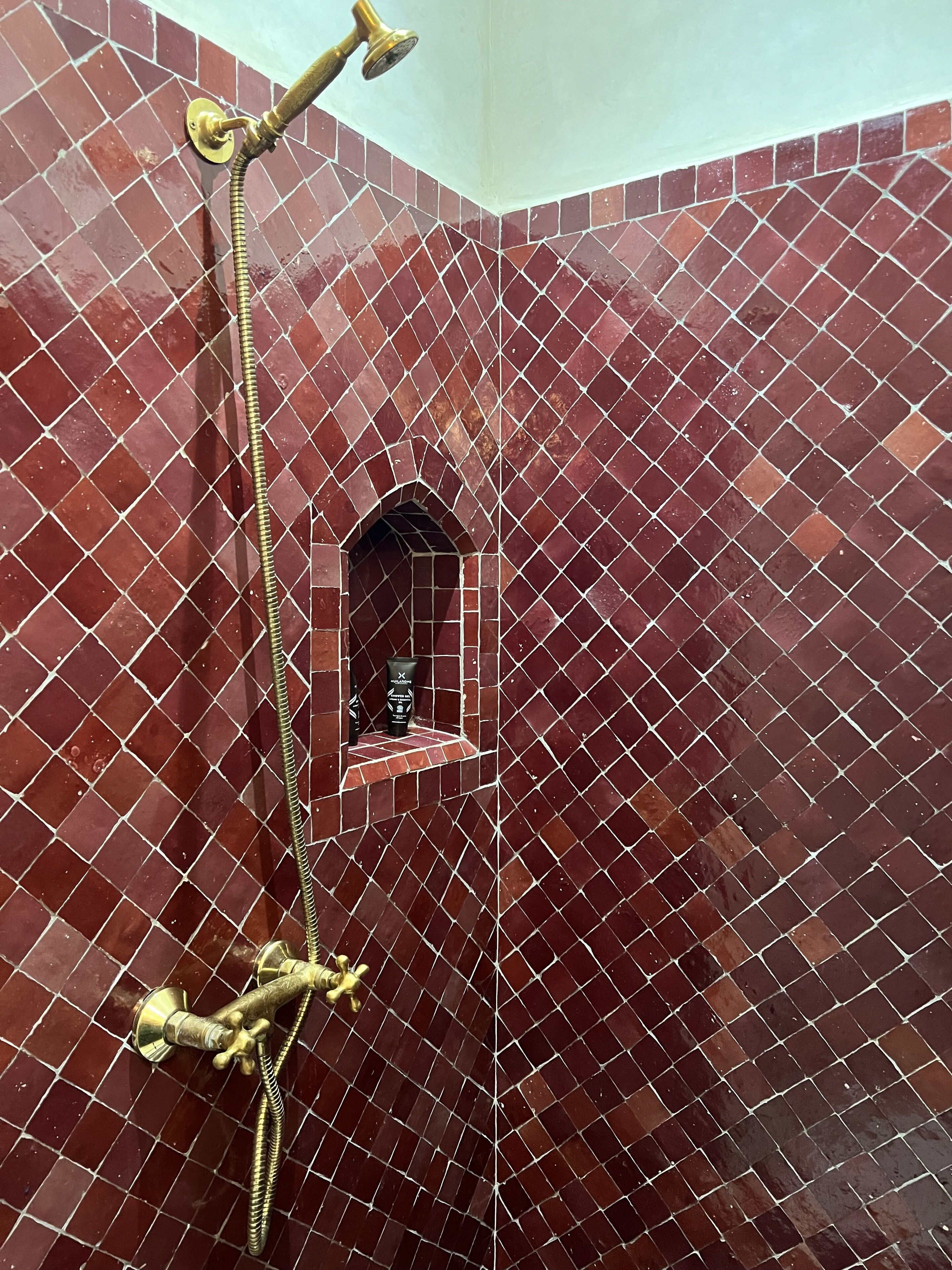 Un área de ducha con pared de azulejos, un cabezal de ducha y un estante con artículos de aseo.
