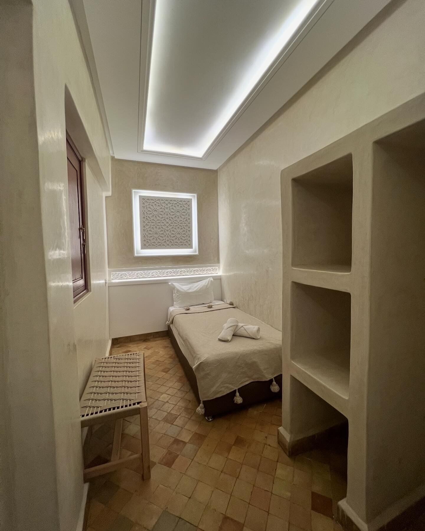 A small, well-lit room with a bed, a window with decorative molding, a shelf unit, and a small table.