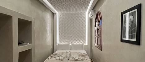 Habitación de hotel estrecha con cama, banco, estantería y alfombra estampada.