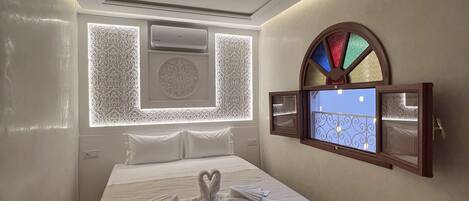 A hotel room with a neatly made bed, a wall-mounted TV, and a decorative window with a stained glass design.