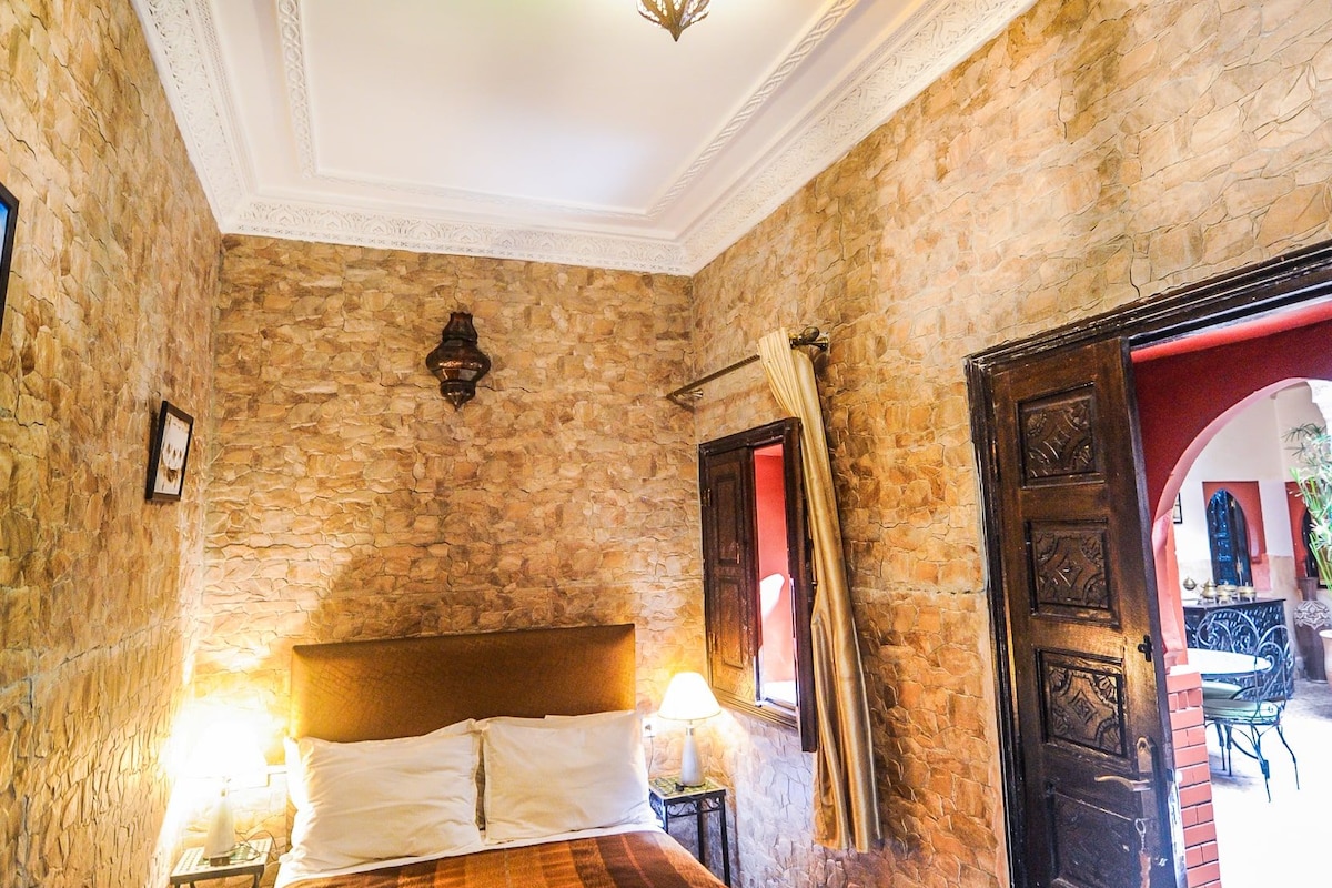 A bedroom with a stone wall, a wooden bed, a chandelier, and a door leading to an outdoor area.