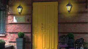 Une porte jaune, deux lampes fixées au mur, un mur de briques, une petite table avec des chaises et une plante en pot.