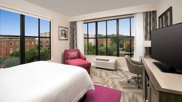 A hotel room with a large bed, a desk with a flat-screen TV, a red armchair, and a large window offering a city view.