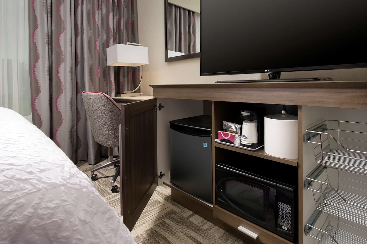 A hotel room with a bed, a desk with a chair, a television, a microwave, a trash bin, and a shelf with toiletries.