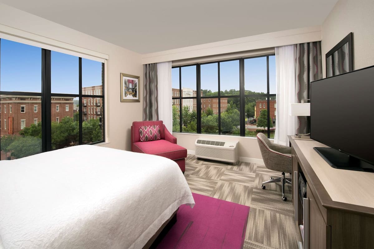 A hotel room with a large bed, a desk with a flat-screen TV, a red armchair, and a large window offering a city view.