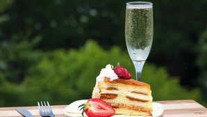 A slice of cake with whipped cream and a strawberry on a plate, accompanied by a glass of champagne, set on a wooden table with a blurred green background. - Hotel Indigo Tuscaloosa Downtown by IHG (Tuscaloosa)
