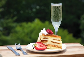 Uma fatia de bolo com chantilly e morango em um prato, acompanhada de uma taça de champanhe, sobre uma mesa de madeira com fundo verde desfocado.