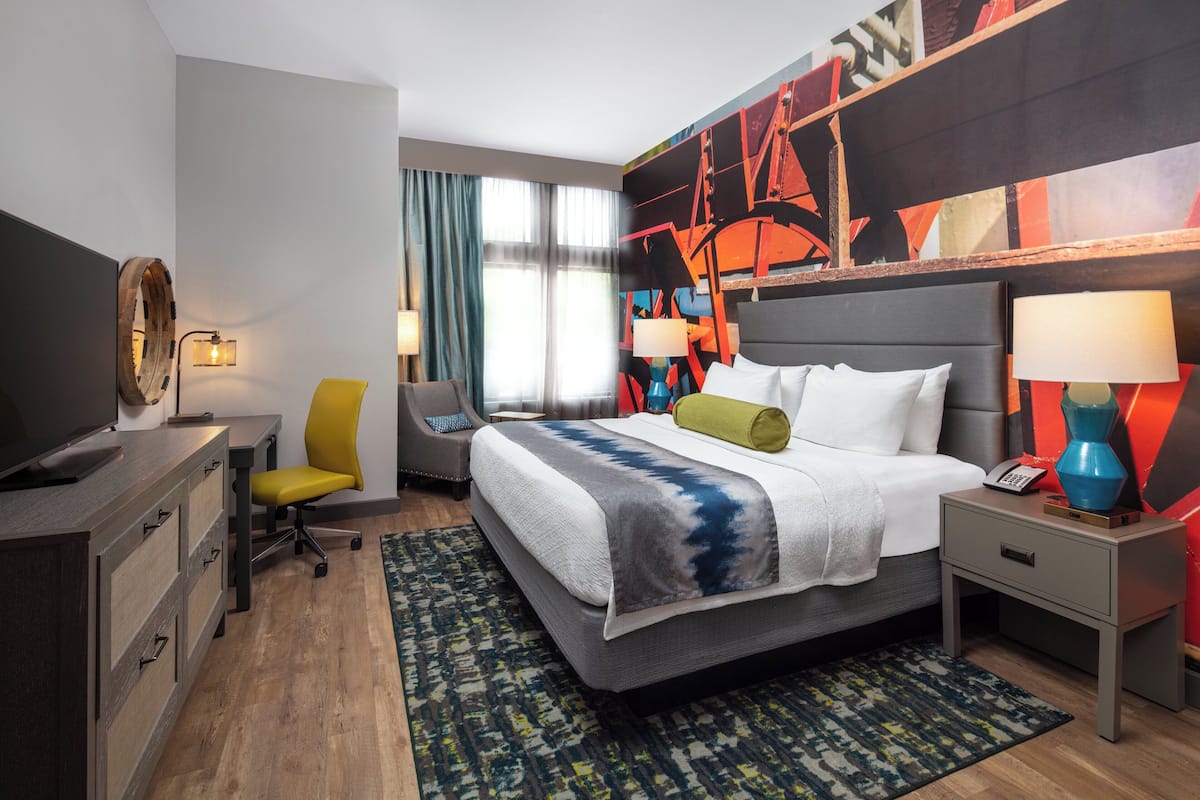 A hotel room with a bed, bedside tables, a desk, a chair, a television, and a large abstract painting on the wall.