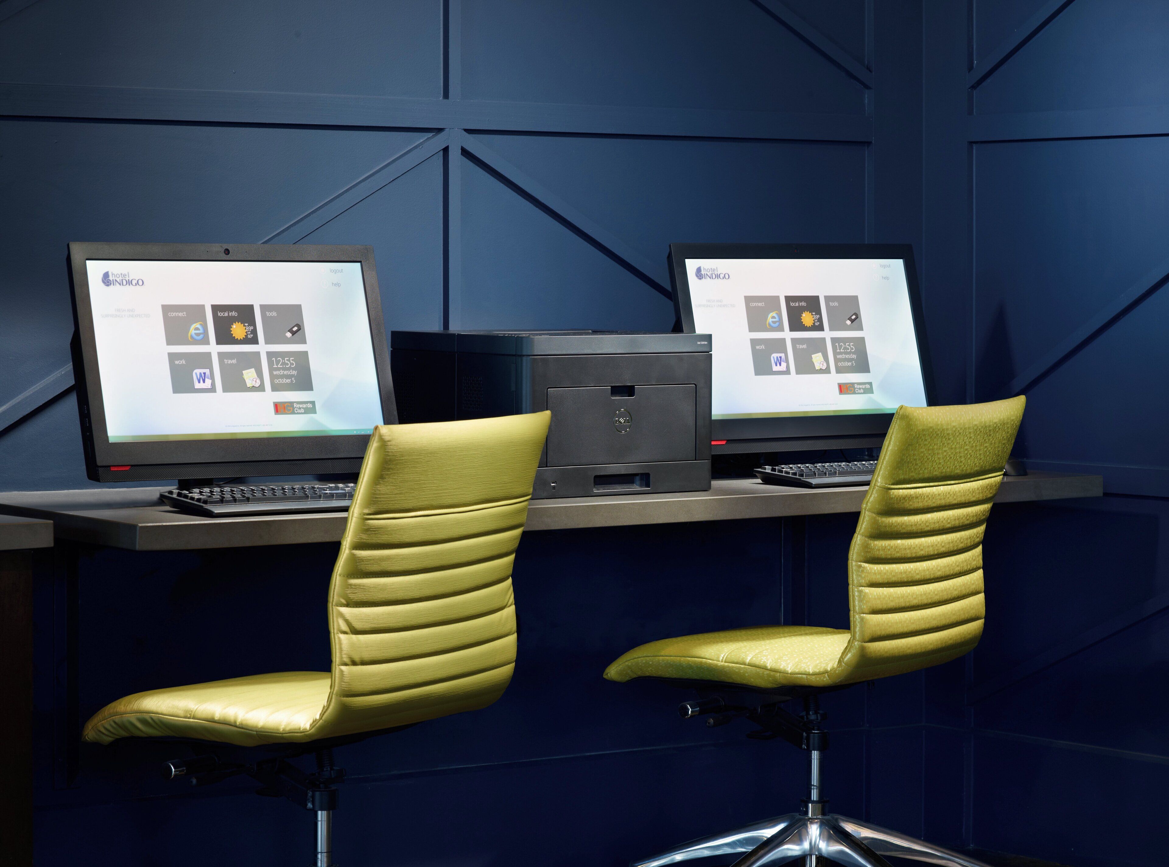 Two computers on a desk with two yellow chairs, a printer, and a wall with a geometric pattern.