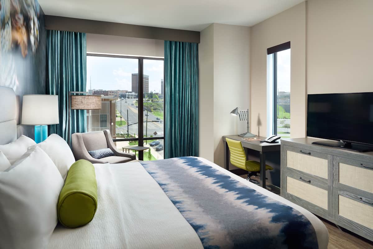 A hotel room with a bed, a desk, a chair, a TV, and a view of the city.