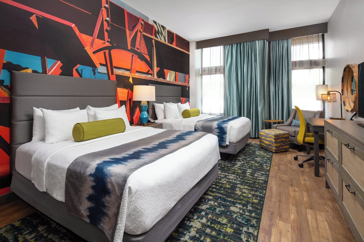 A hotel room with two beds, a desk, a chair, and a large abstract painting on the wall.